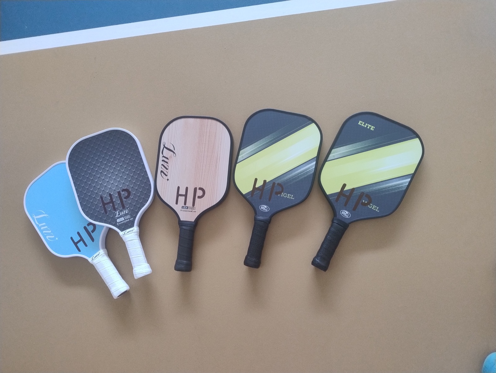 Five pickleball paddles of different shapes, face sizes and grip styles arranged on an indoor court