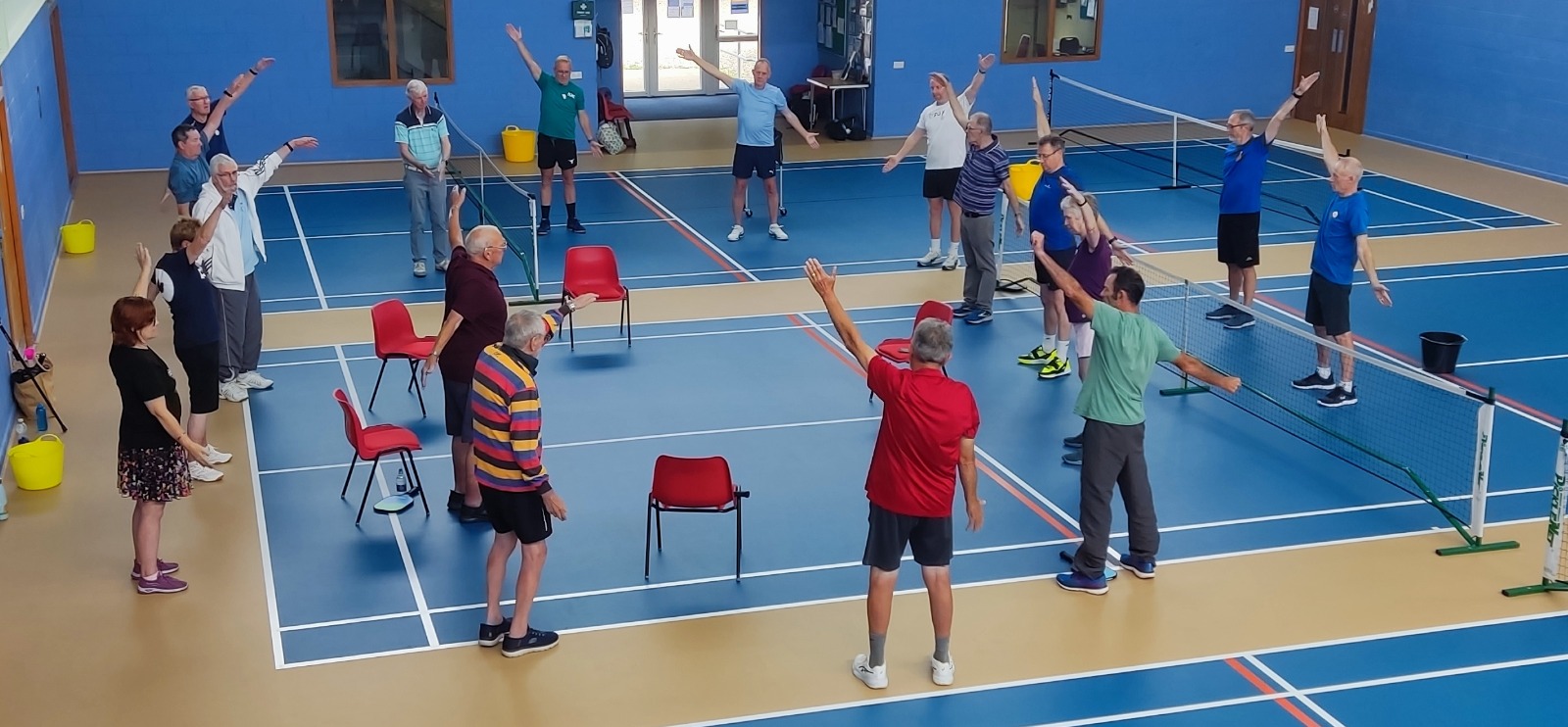 Players and volunteers performing a standing group warm-up on an indoor pickleball court, with chairs available nearby