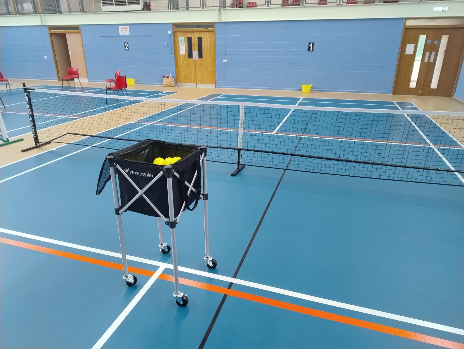 A raised wheeled basket containing pickleballs beside an indoor pickleball court