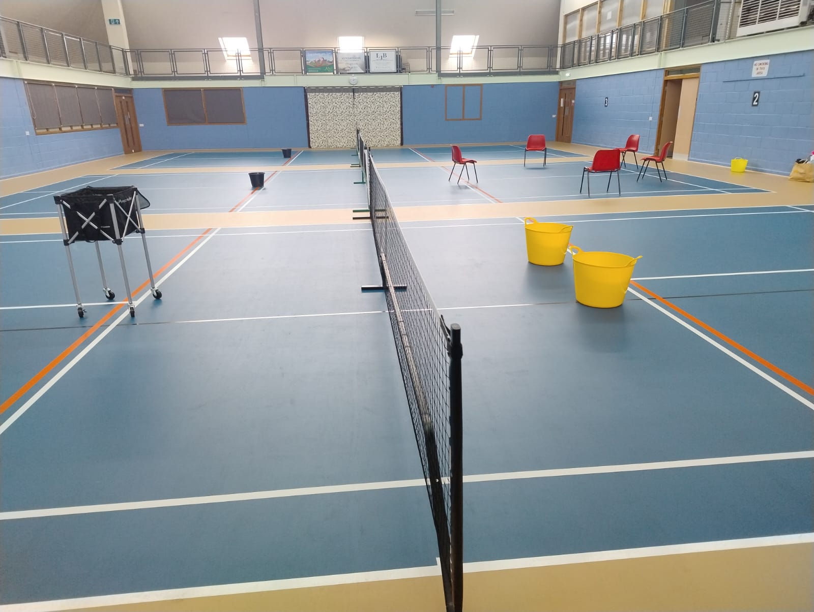 Three indoor pickleball courts prepared with ball baskets, flexible buckets and red chairs for a group warm-up